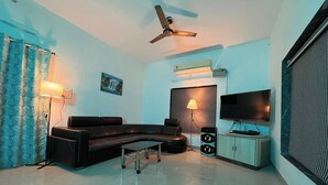 A modern living room with a sectional sofa, a flat-screen TV, ceiling fan, and a sound system. - Lsq Sunita Farms (Karjat)