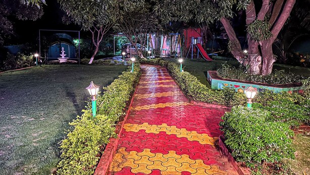 A nighttime garden pathway with a red and yellow patterned walkway, surrounded by greenery and trees. - Lsq Sunita Farms (Karjat)