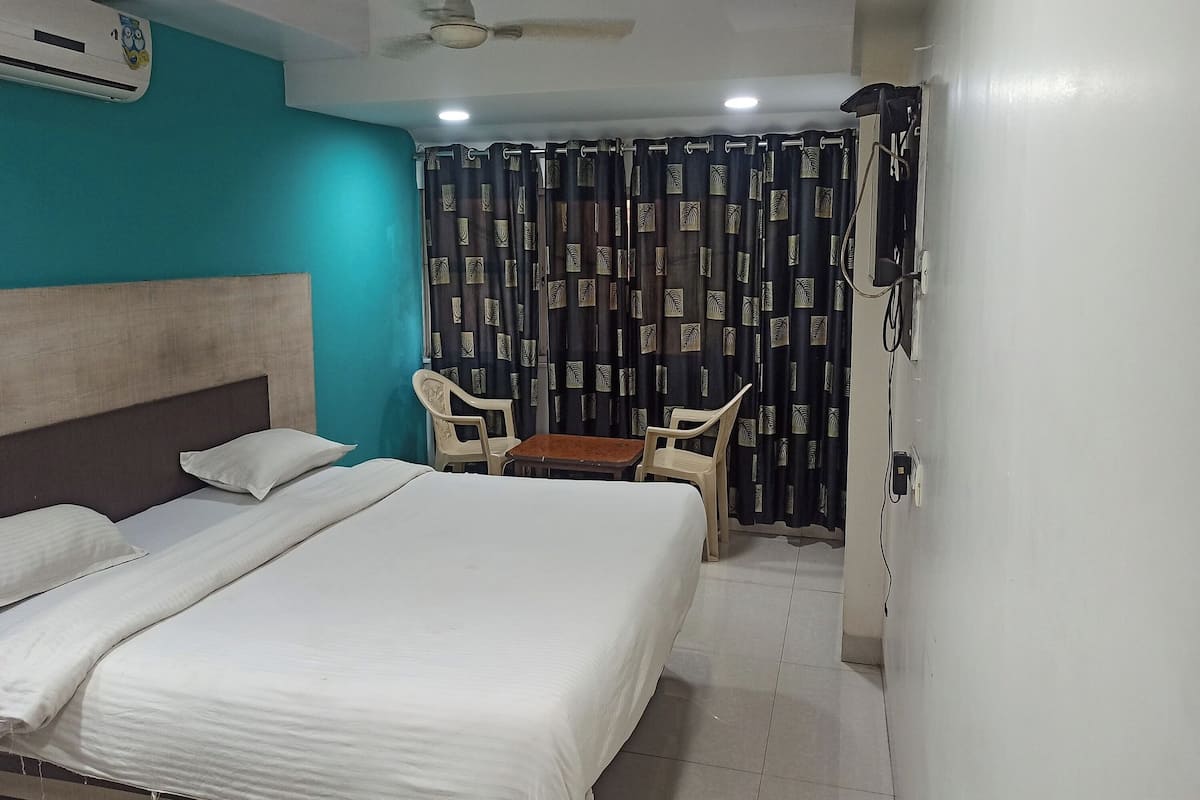 A hotel room with a bed, a desk, two chairs, a ceiling fan, and a curtain with a pattern.