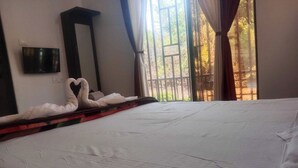 A neatly made bed with a heart-shaped towel arrangement, a flat-screen TV mounted on the wall, and a window with curtains. - Devansh Villa (Mahabaleshwar)