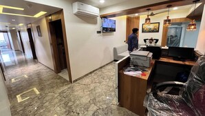 Zona de recepción de un hotel con piso de mármol, mostrador de recepción, un televisor y un panel de pared con marco de madera.