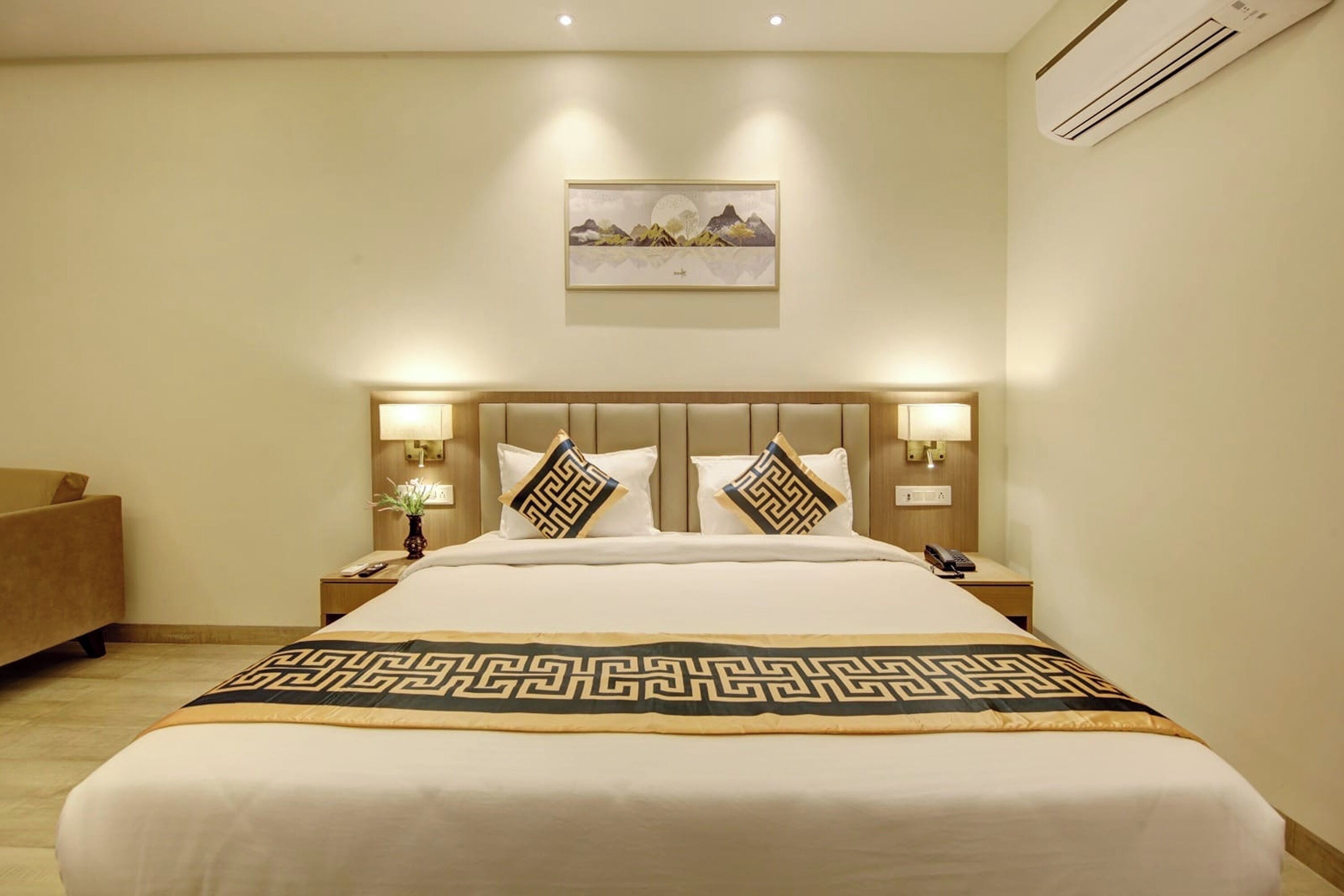A hotel room with a large bed, two bedside tables, a nightstand, a lamp, a painting on the wall, and an air conditioning unit.