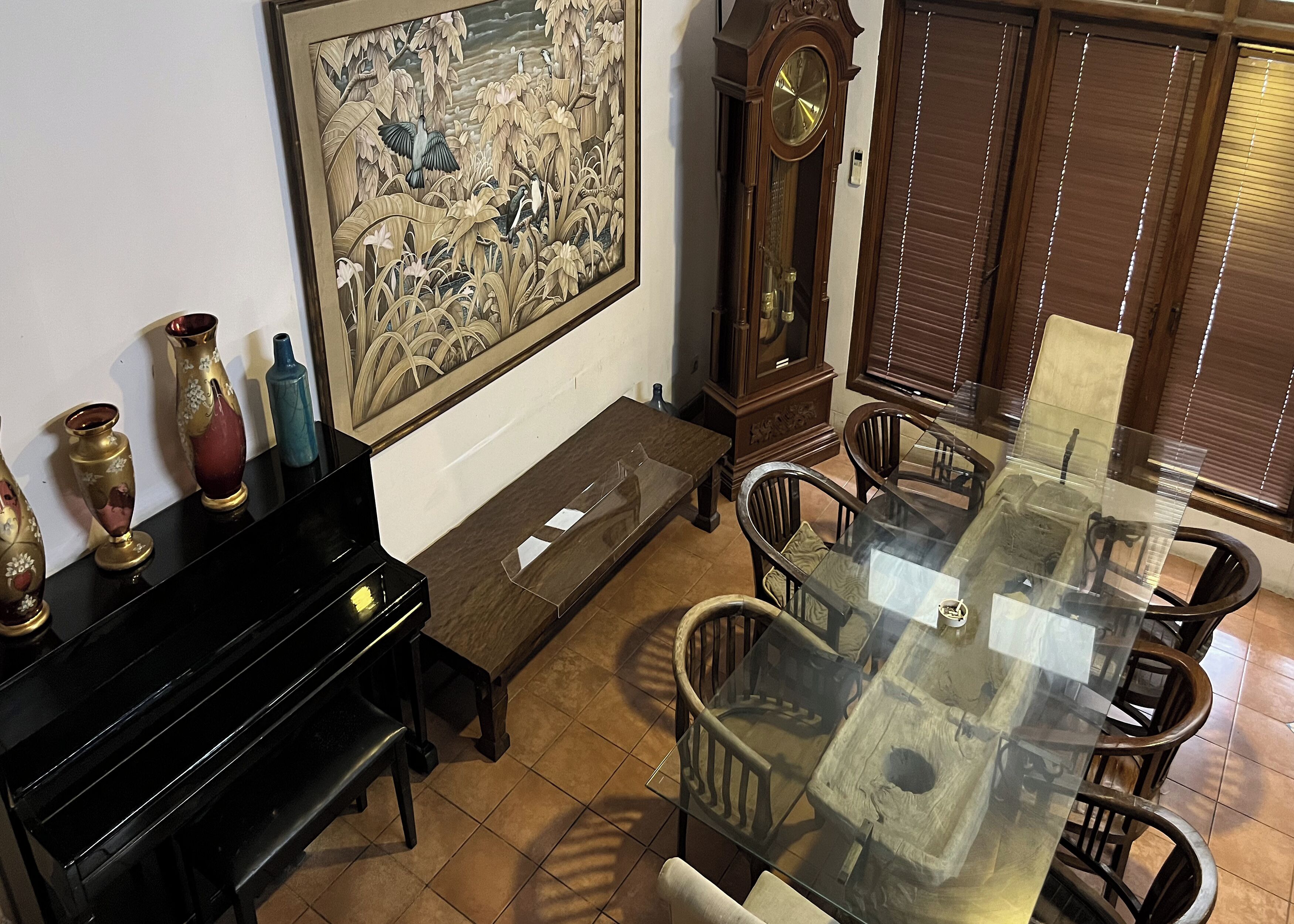 A dining area with a glass-top table, wooden chairs, a piano, and a large framed painting on the wall.