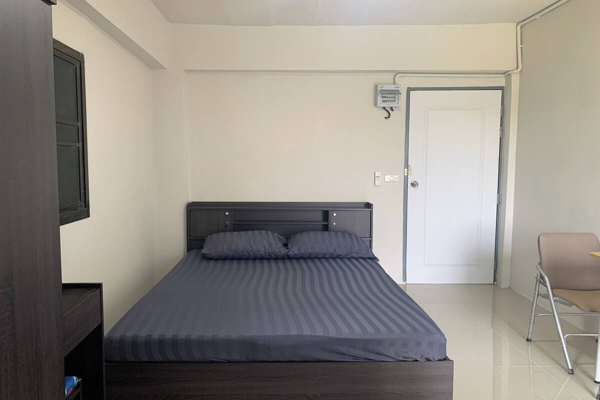 Un dormitorio con cama, una silla, una puerta y un cuadro eléctrico en la pared.
