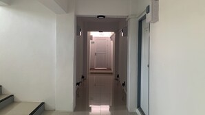 A hallway with a door at the end, a light fixture on the ceiling, and a wall-mounted control panel. - Na Nont Place (Pak Kret)