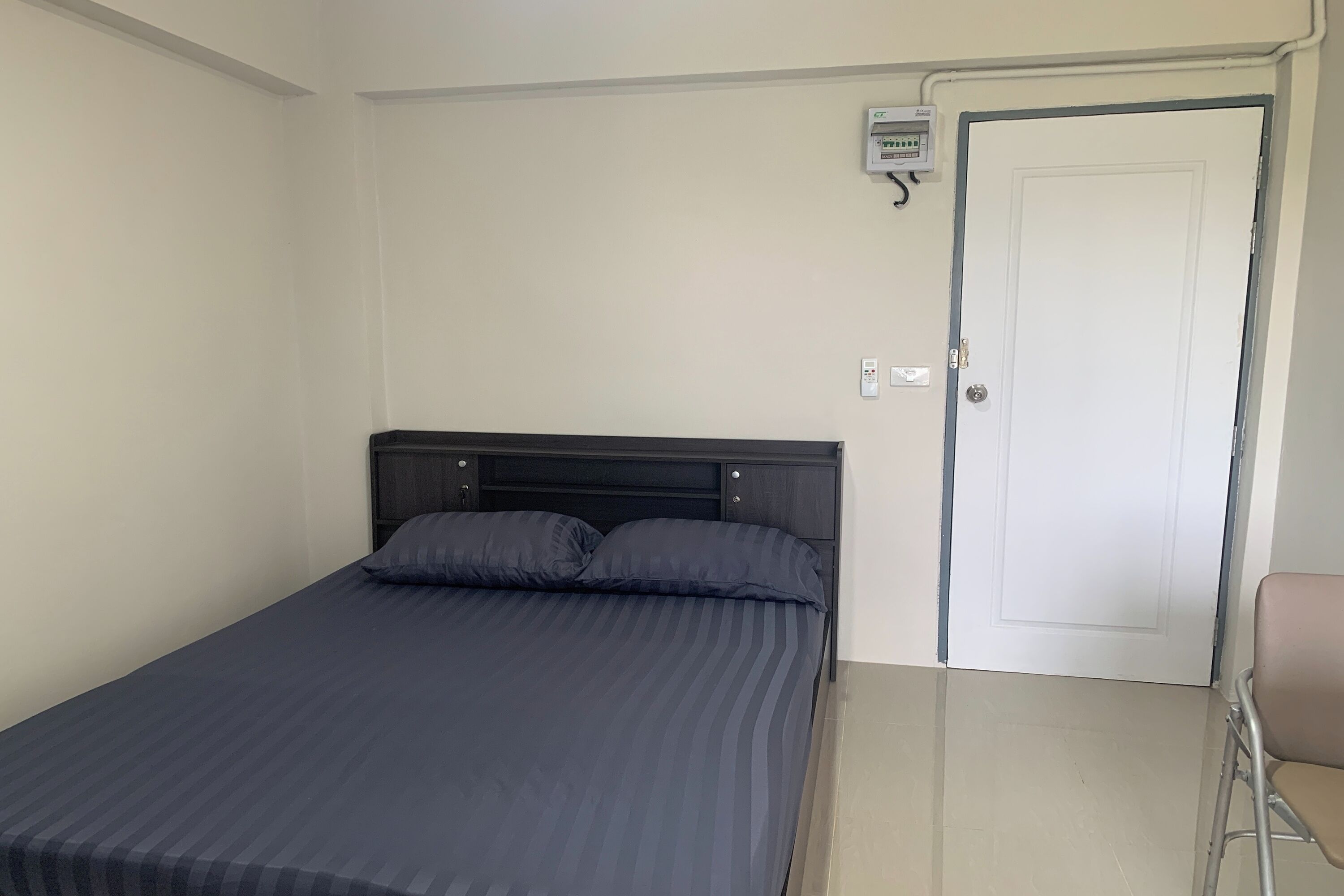 Un letto singolo con una testata scura e due cuscini, una porta bianca con maniglia e una piccola presa elettrica sulla parete.