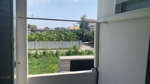 A view through a window with an air conditioning unit, a metal fence, and greenery in the background. - Na Nont Place (Pak Kret)