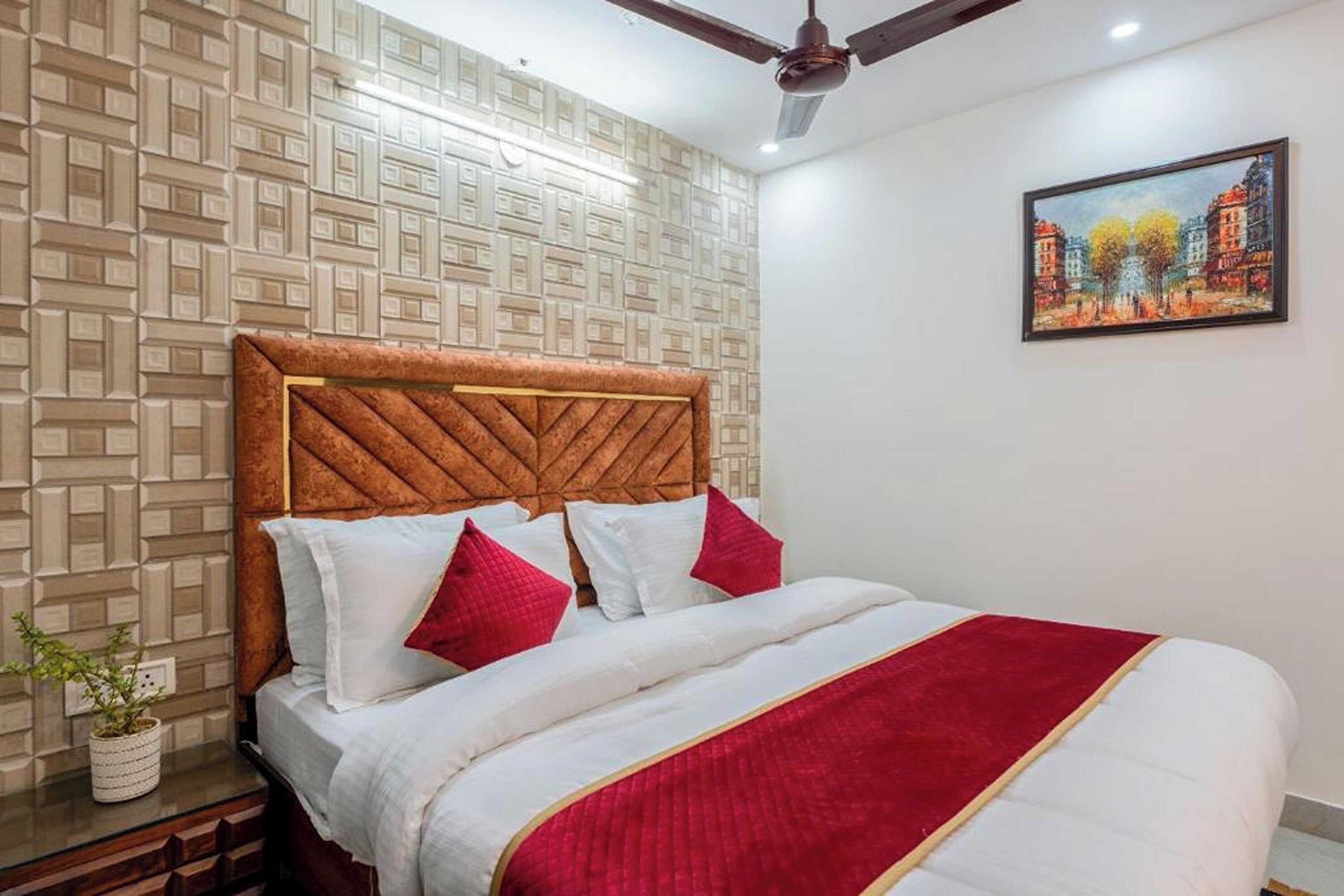 Una habitación de hotel con pared estampada, cabecera de madera, cama con ropa de cama blanca y detalles rojos, un cuadro en la pared y un ventilador de techo.