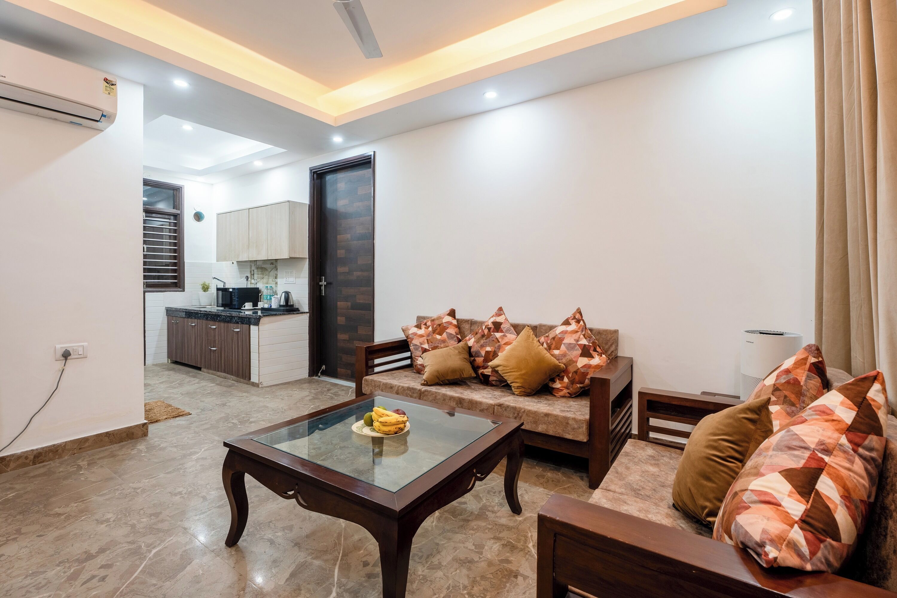 A modern living room with a glass coffee table, a sofa with patterned cushions, and a kitchen in the background.