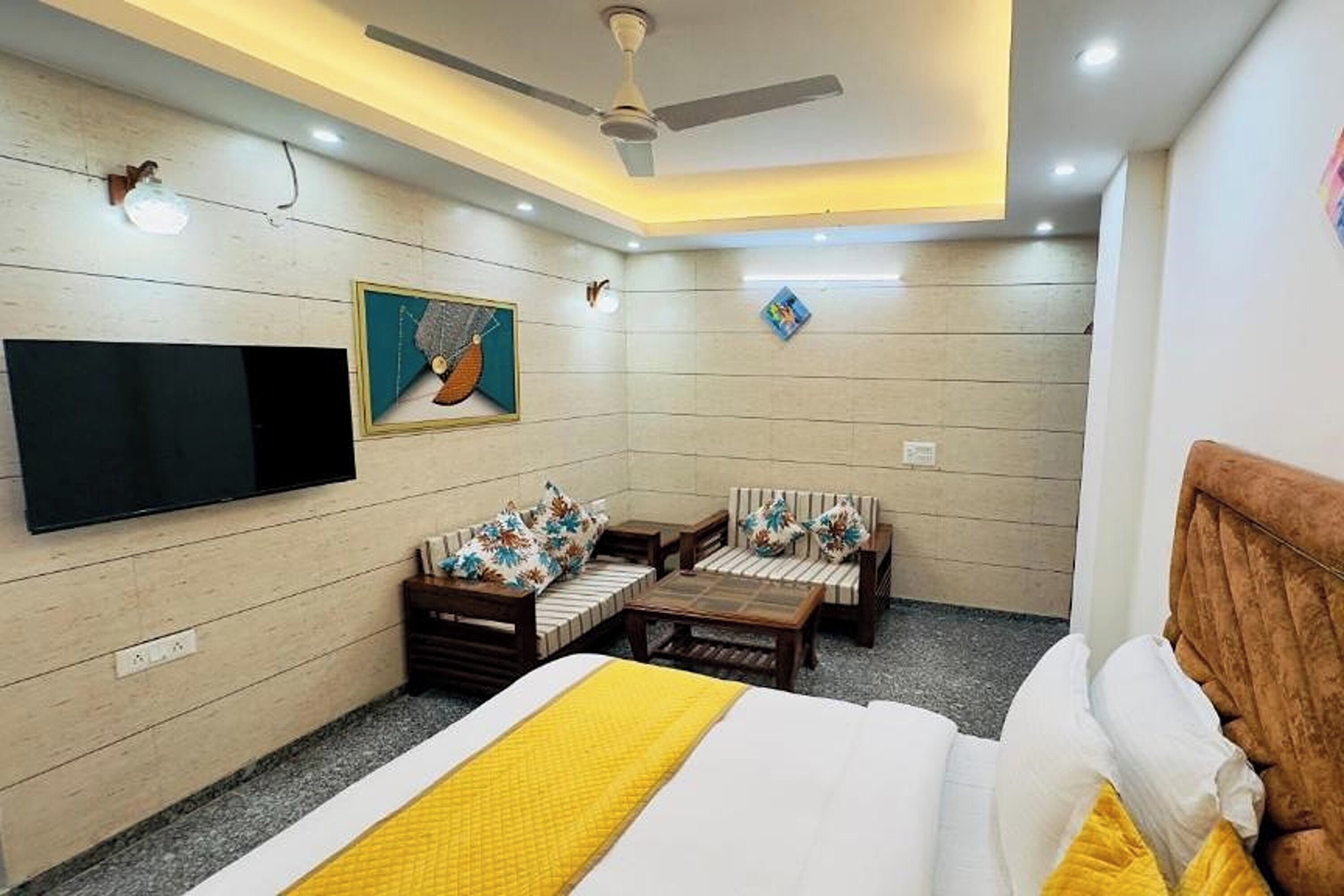 A hotel room with a bed, a sofa, a coffee table, a TV mounted on the wall, and a ceiling fan.