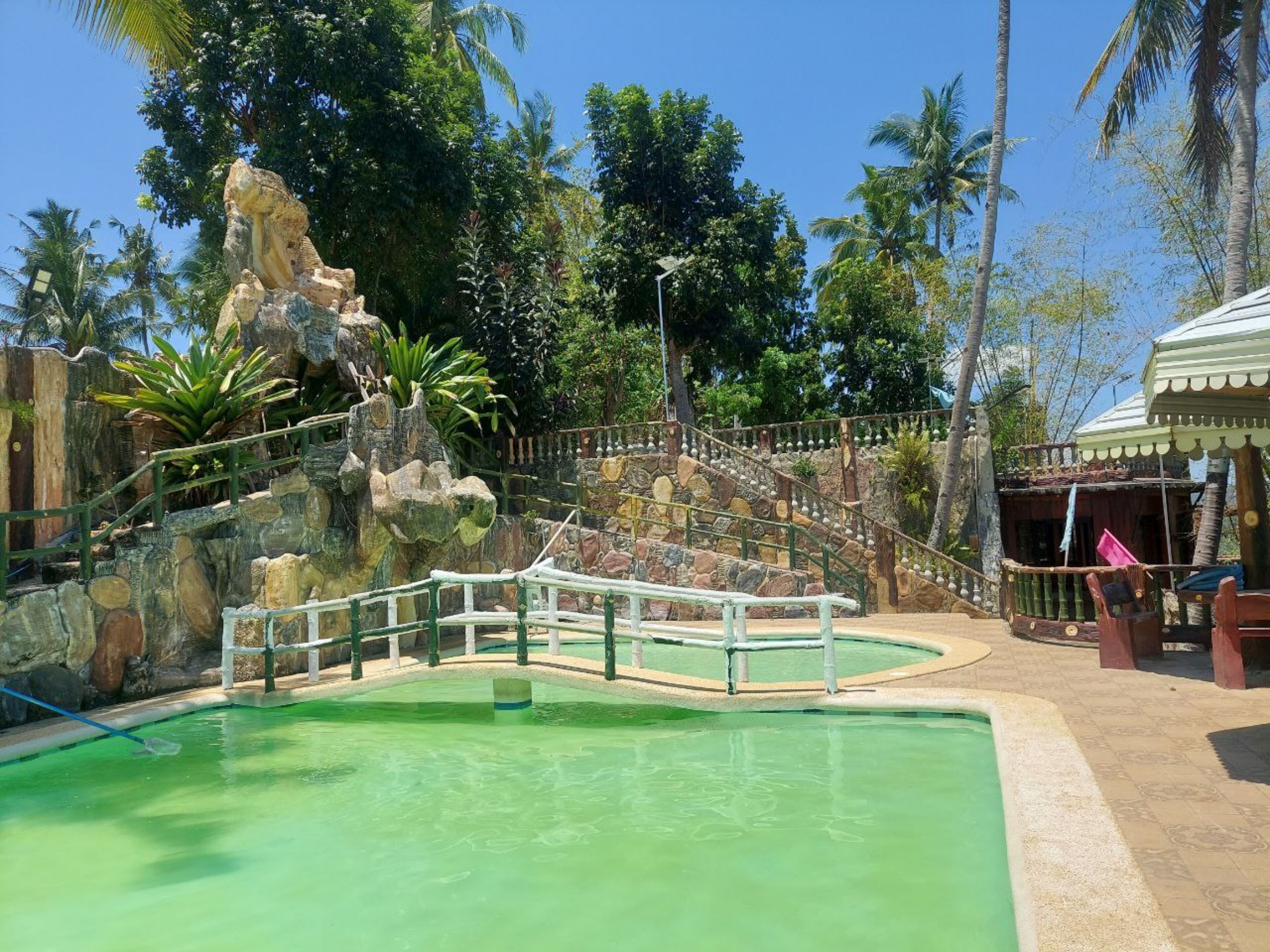 A pool area with a stone waterfall feature, surrounded by palm trees and a wooden fence.