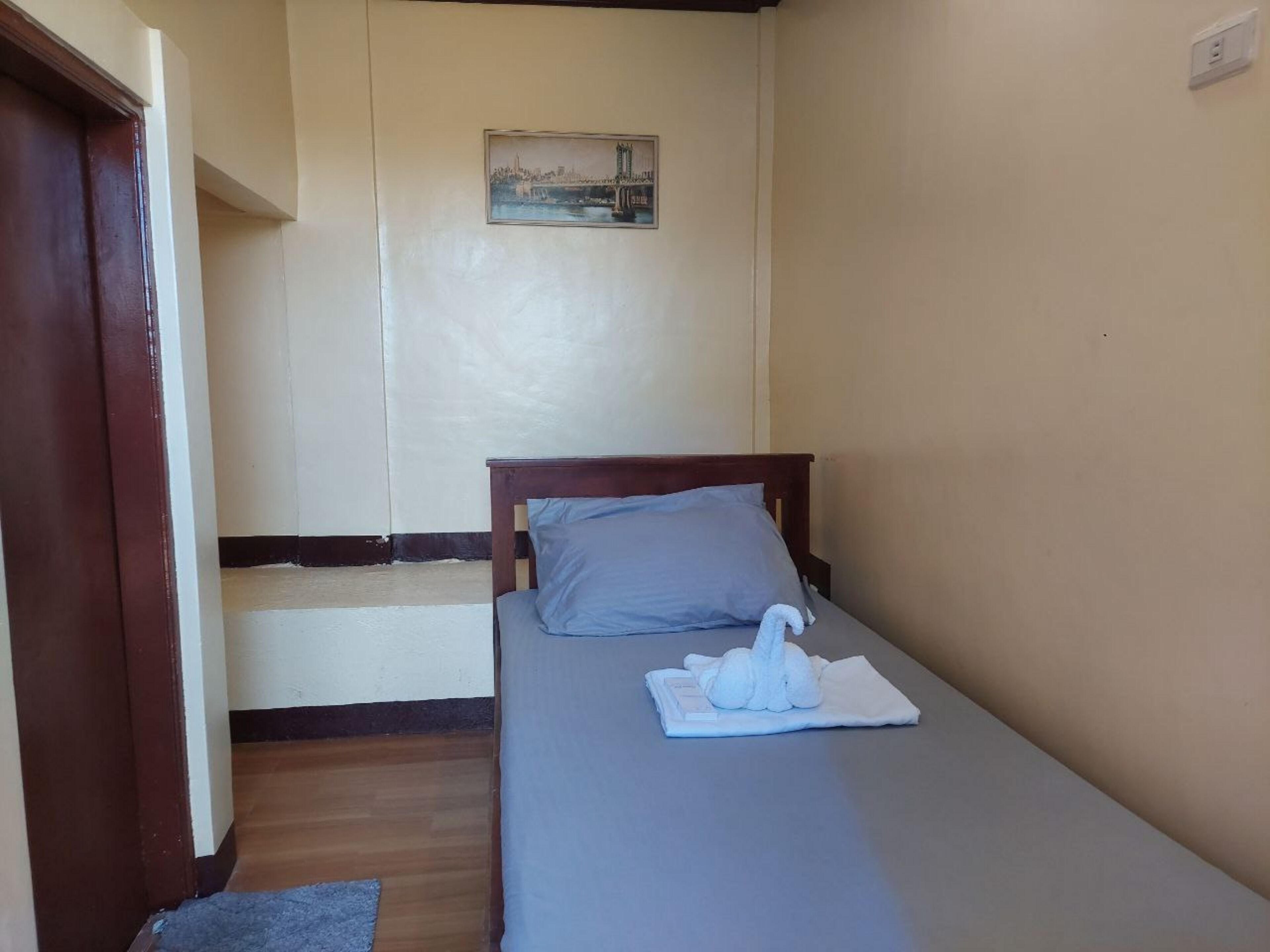 Una habitación pequeña con cama individual, cabecera de madera, almohada gris y toalla blanca sobre la cama. Se observa un cuadro enmarcado en la pared y una puerta de madera a la izquierda.