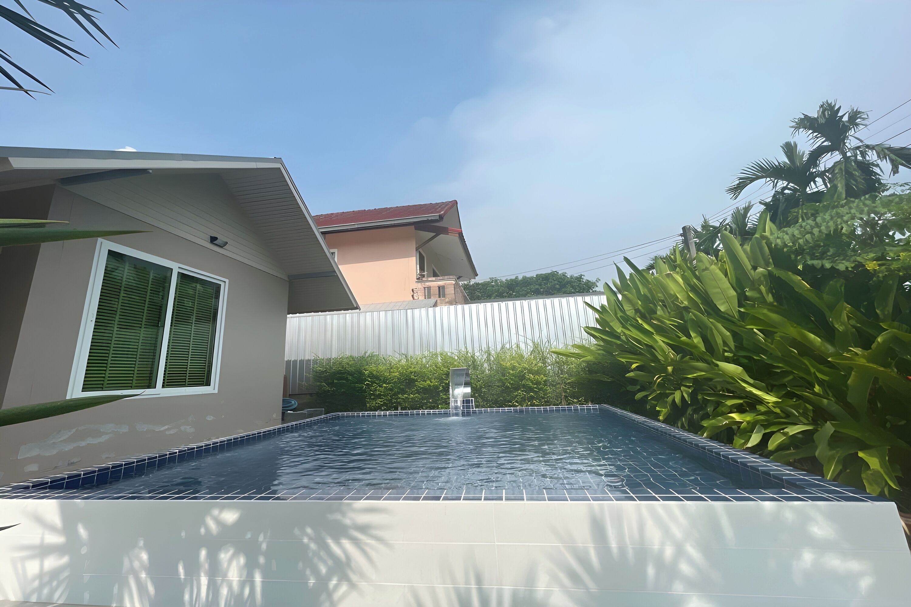 Una piscina con borde de azulejos, rodeada de vegetación y un edificio de techo rojo.