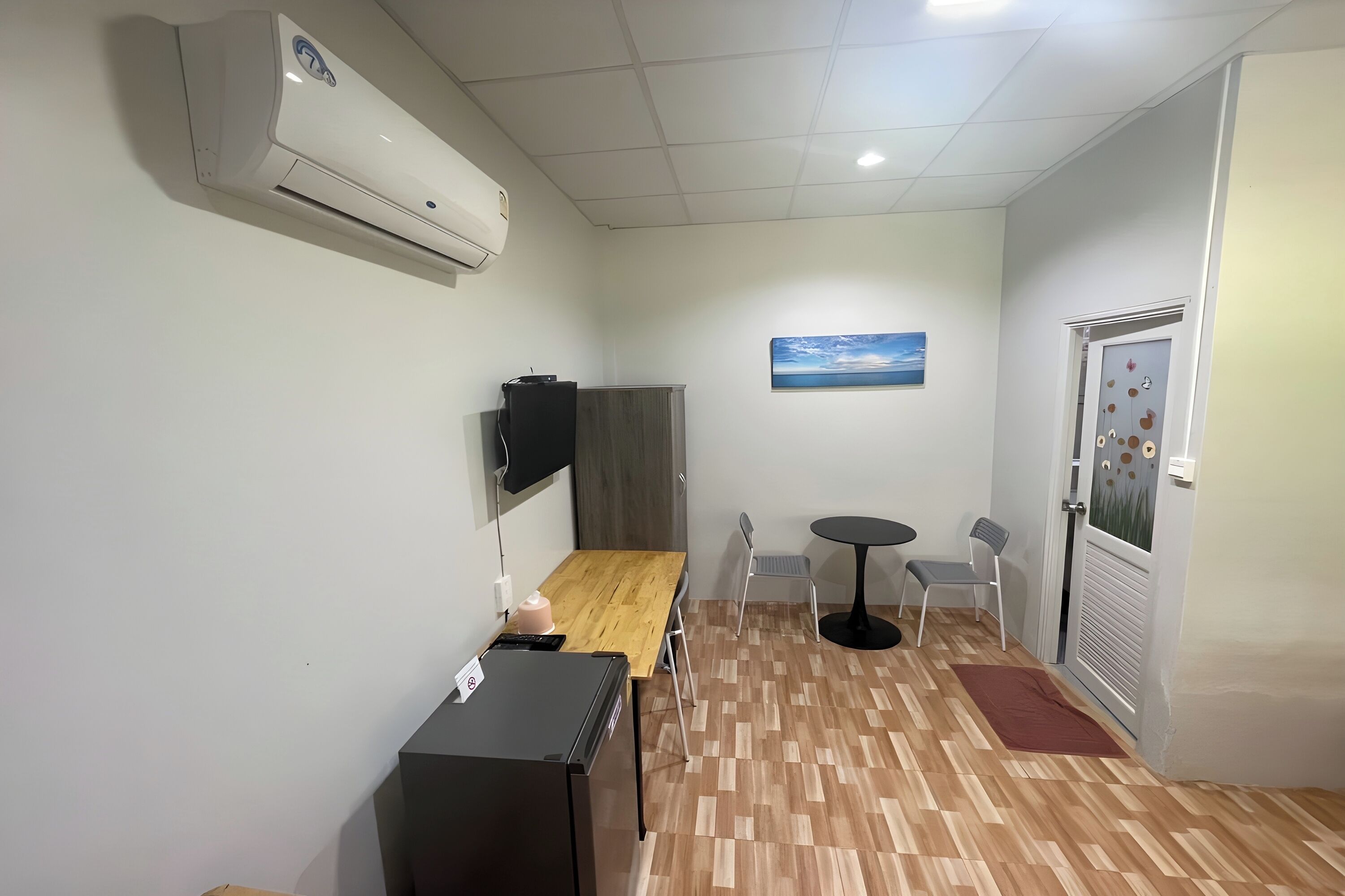 Una habitación pequeña y limpia con piso de madera, una cama, un escritorio, una silla, una mesita y una puerta con ventana.