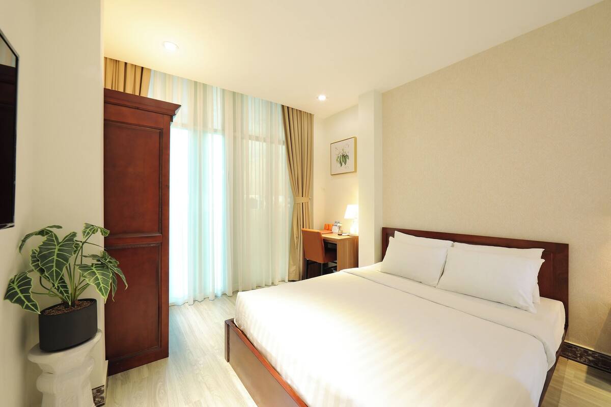 Une chambre d’hôtel avec un grand lit, un bureau, une chaise, une plante en pot et une armoire en bois.