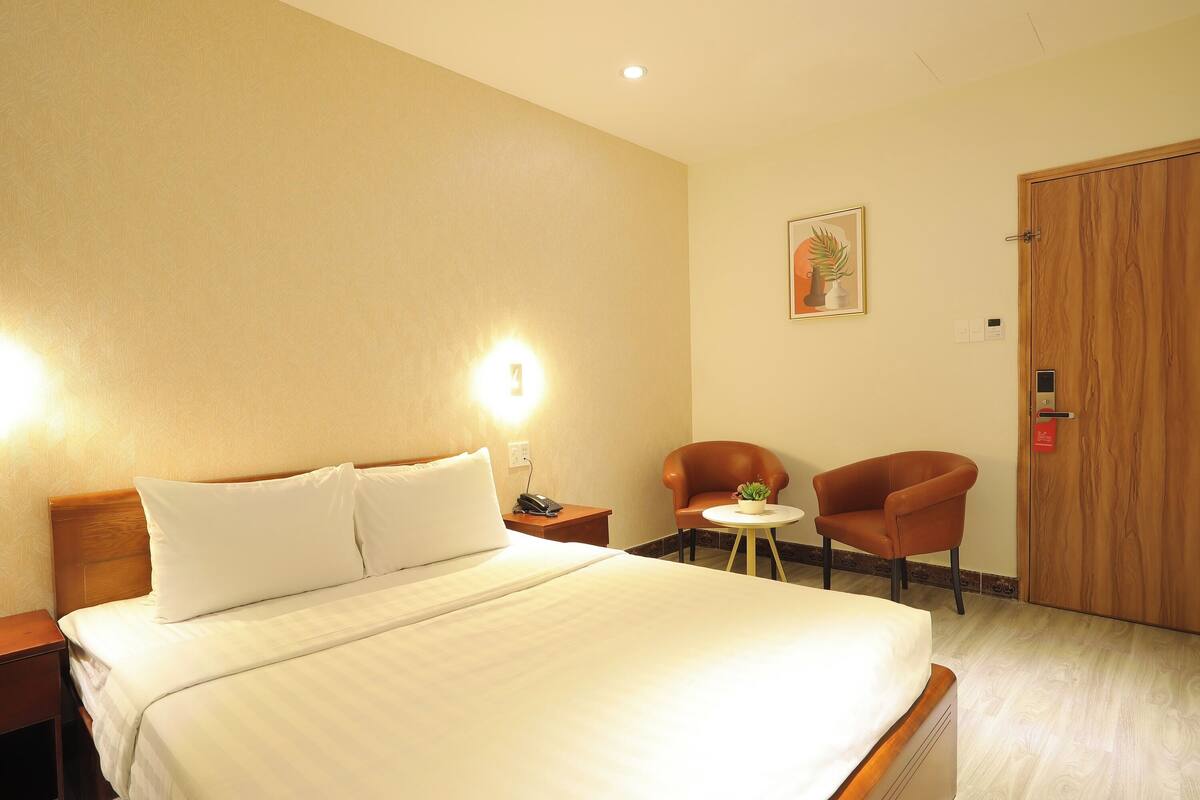 Une chambre d’hôtel avec un grand lit, deux chaises, une petite table avec une plante et une porte en bois.