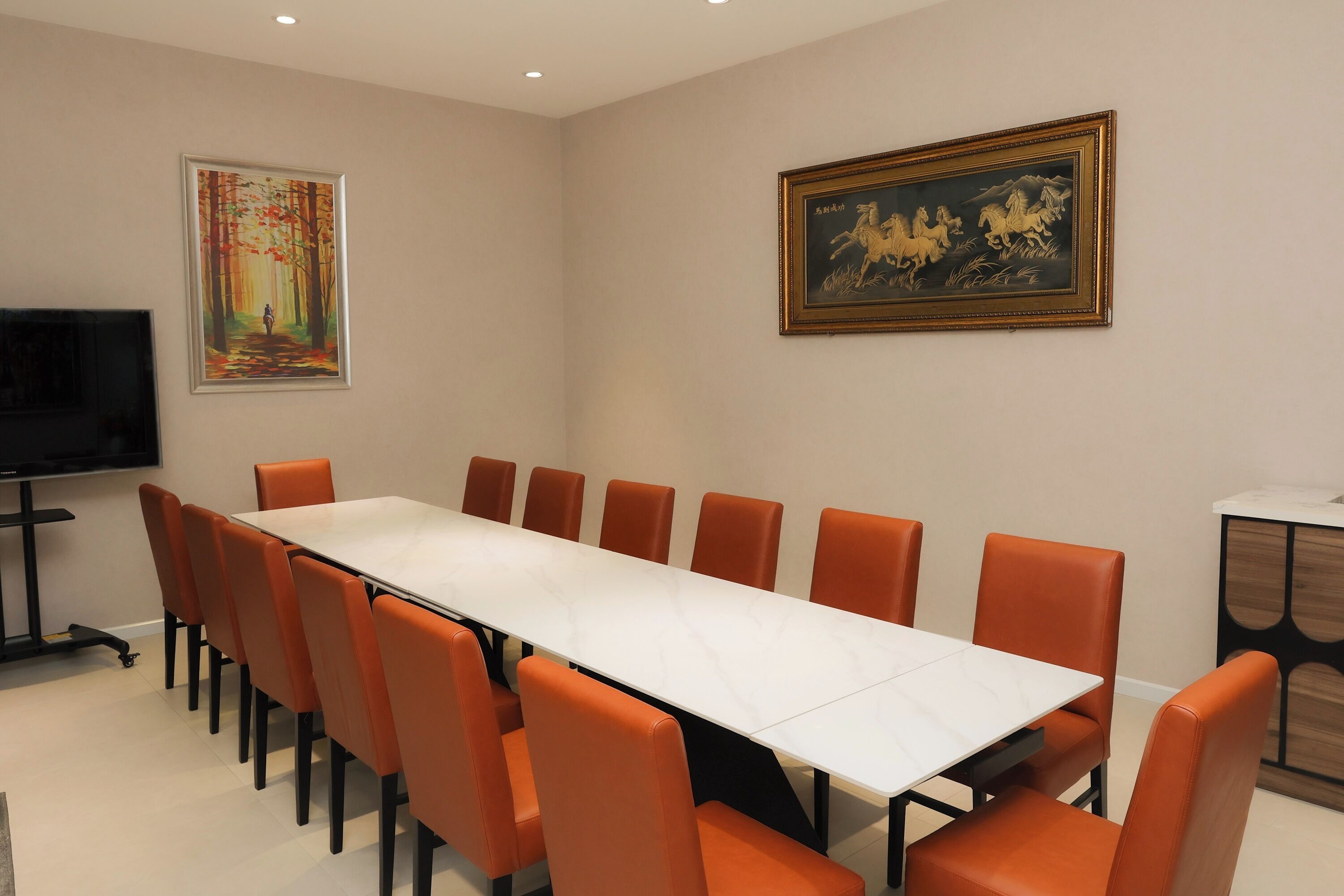 Un salón de conferencias con una larga mesa y sillas color naranja, un televisor de pantalla plana y dos cuadros enmarcados en las paredes.