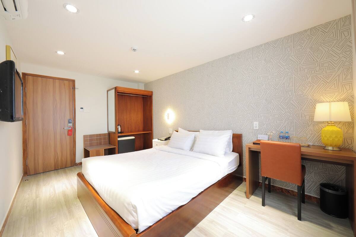 Une chambre d’hôtel avec un grand lit, un bureau avec une chaise, une lampe de bureau et un mur décoré d’un motif.