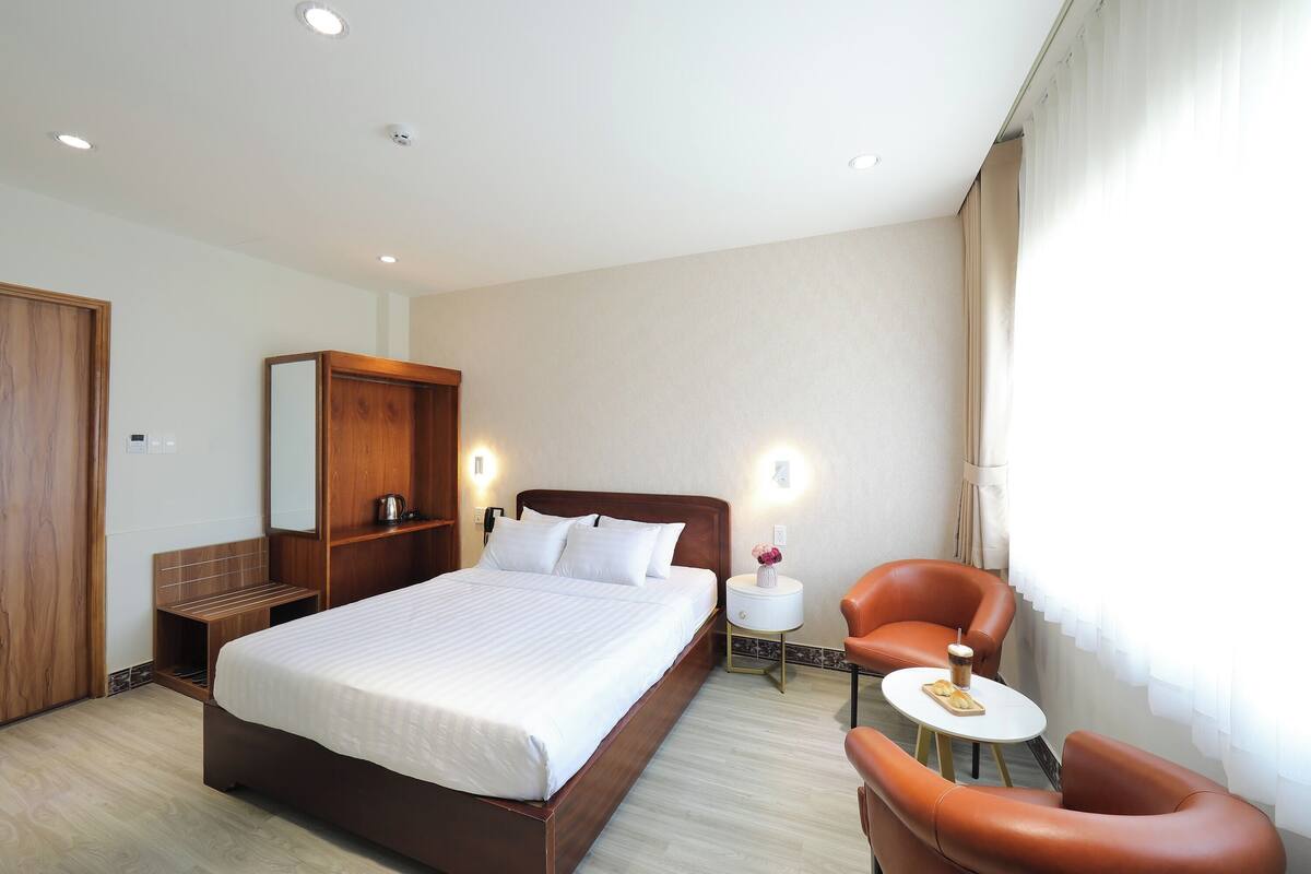 Une chambre d’hôtel avec un grand lit, une armoire en bois, une petite table d’appoint, une chaise et une fenêtre avec des rideaux.