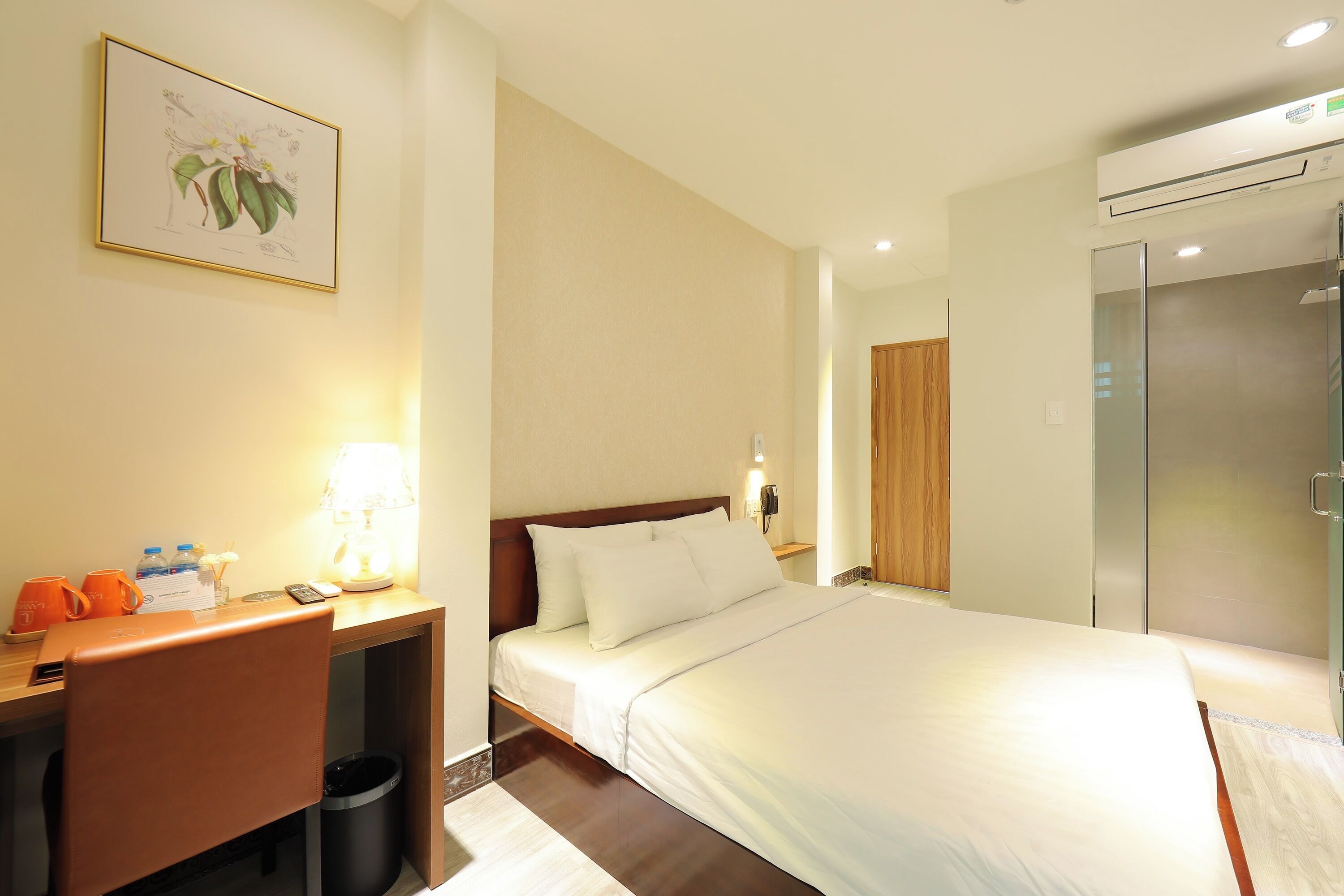 A hotel room with a bed, desk, chair, lamp, and a framed botanical illustration on the wall.
