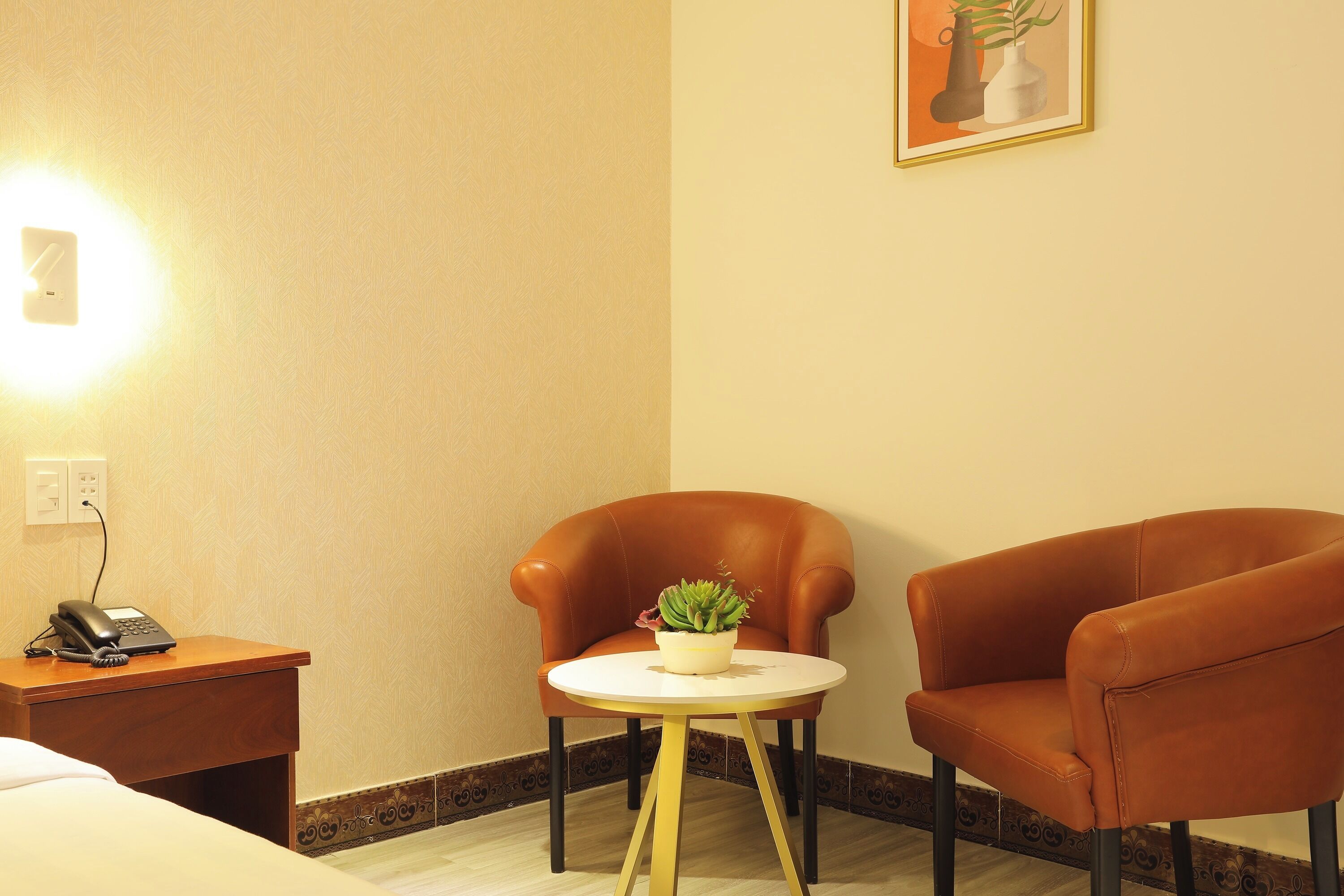 A hotel room with a bed, a desk with a phone, a small round table with a plant, and two armchairs.