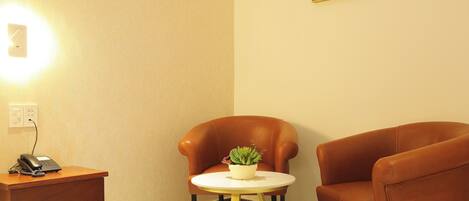 A hotel room with a bed, a desk with a phone, a small round table with a plant, and two armchairs.