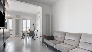 A modern living room with a sofa, a dining area with chairs and a table, a kitchen area with a TV, and a radiator.