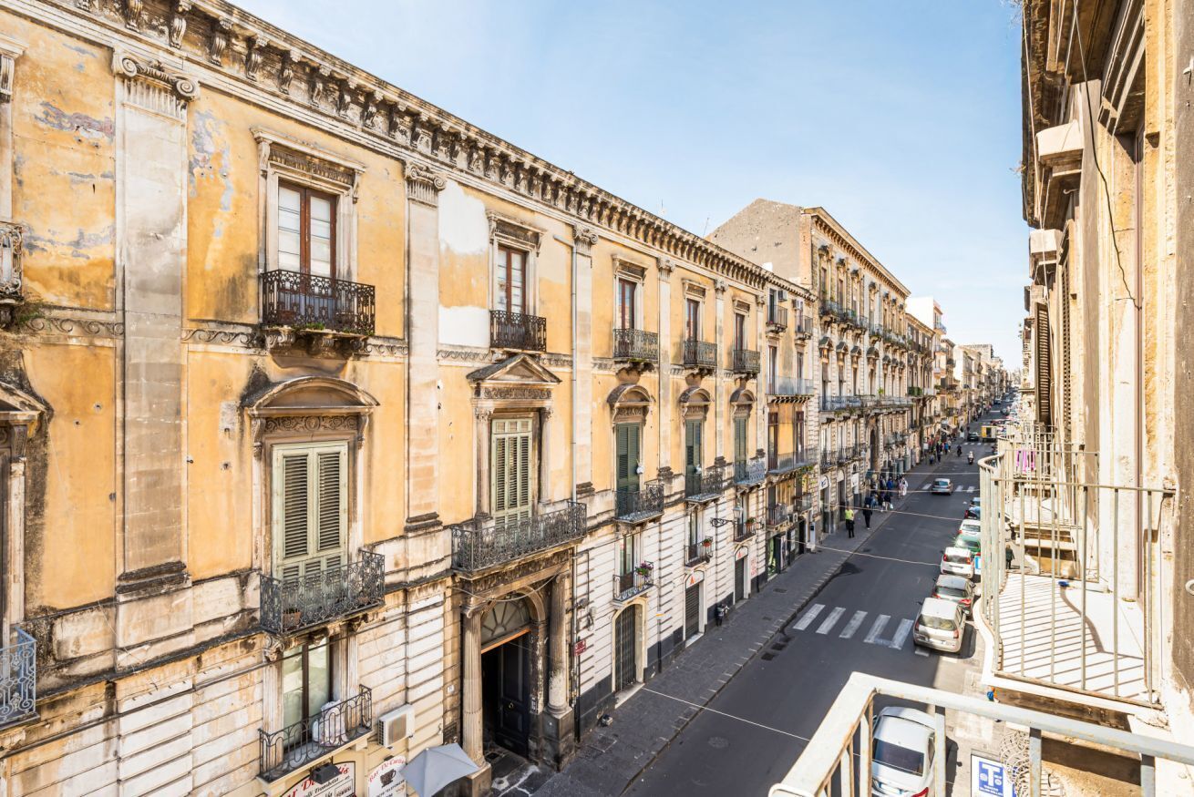 A row of historic buildings with balconies, a clear street, and parked cars.