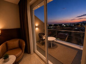 Ein Balkon mit Blick auf eine Stadtlandschaft im Abenddämmerung, ein Ledersofa, ein kleiner Tisch mit einer Pflanze und ein Korbstuhl.