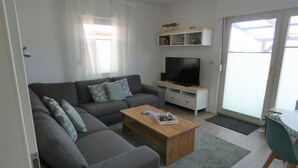 A modern living room with a grey sofa, a wooden coffee table, a flat-screen TV on a white stand, and a sliding glass door leading to an outdoor area. - Wellmann Modern Retreat (Borkum)