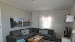 A modern living room with a grey sofa, a wooden coffee table, and a dining table with green chairs. There are two wall-mounted paintings and a TV on a small table. - Wellmann Modern Retreat (Borkum)