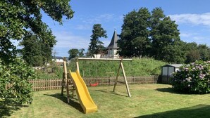 A playground with a yellow slide and a swing set in a grassy yard with a wooden fence and trees. - Wellmann Modern Retreat (Borkum)