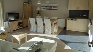 Una sala de estar moderna con televisor, mesa de comedor y sillas. Se observa un área de cocina con armarios y un lavamanos.