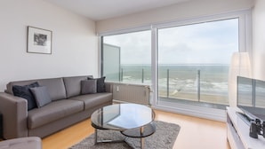 A modern living room with a sofa, a glass coffee table, and a large window offering a view of the sea. - Cozy 1-bedroom Apartment With Sea View (Middelkerke)