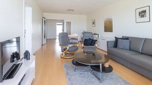 A modern living room with a flat-screen TV, a sofa, a coffee table, and a dining area with chairs. - Cozy 1-bedroom Apartment With Sea View (Middelkerke)