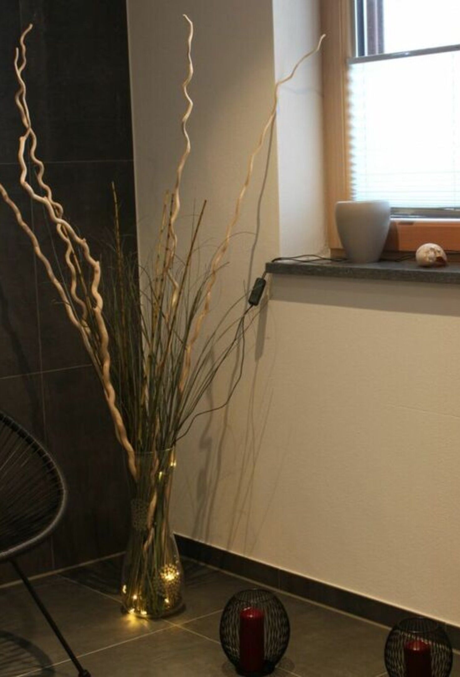 A modern interior with a vase of branches, a chair, and a window with blinds.
