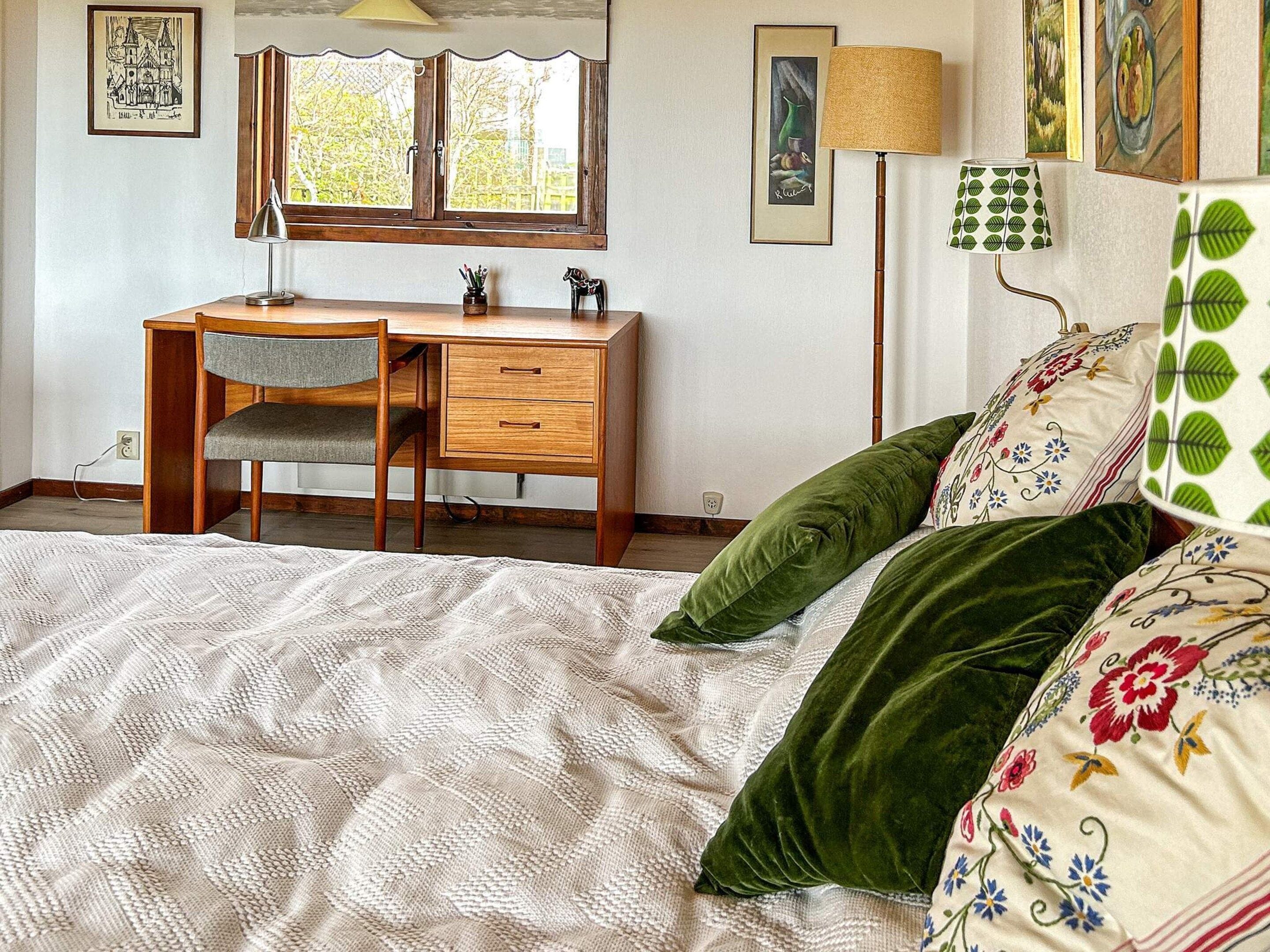 A bedroom with a bed, a desk, and a chair. There are two windows, one with a view of trees and another with a view of a building.