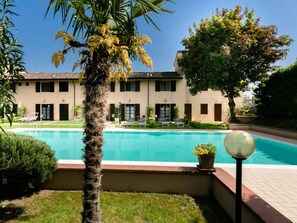 A pool area with a palm tree, a building with a tiled roof, and a clear blue sky. - Casa Vecchio Mulino 1 (Peschiera del Garda)