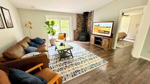 Een woonkamer met een bruine leren bank, een salontafel, een televisie op een houten standaard, een open haard en een groot raam.