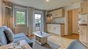 A modern living room with a sofa, a coffee table, and a dining table. There is a kitchen area with cabinets and a window. - Haus Leutner Bodenmais (Bodenmais)