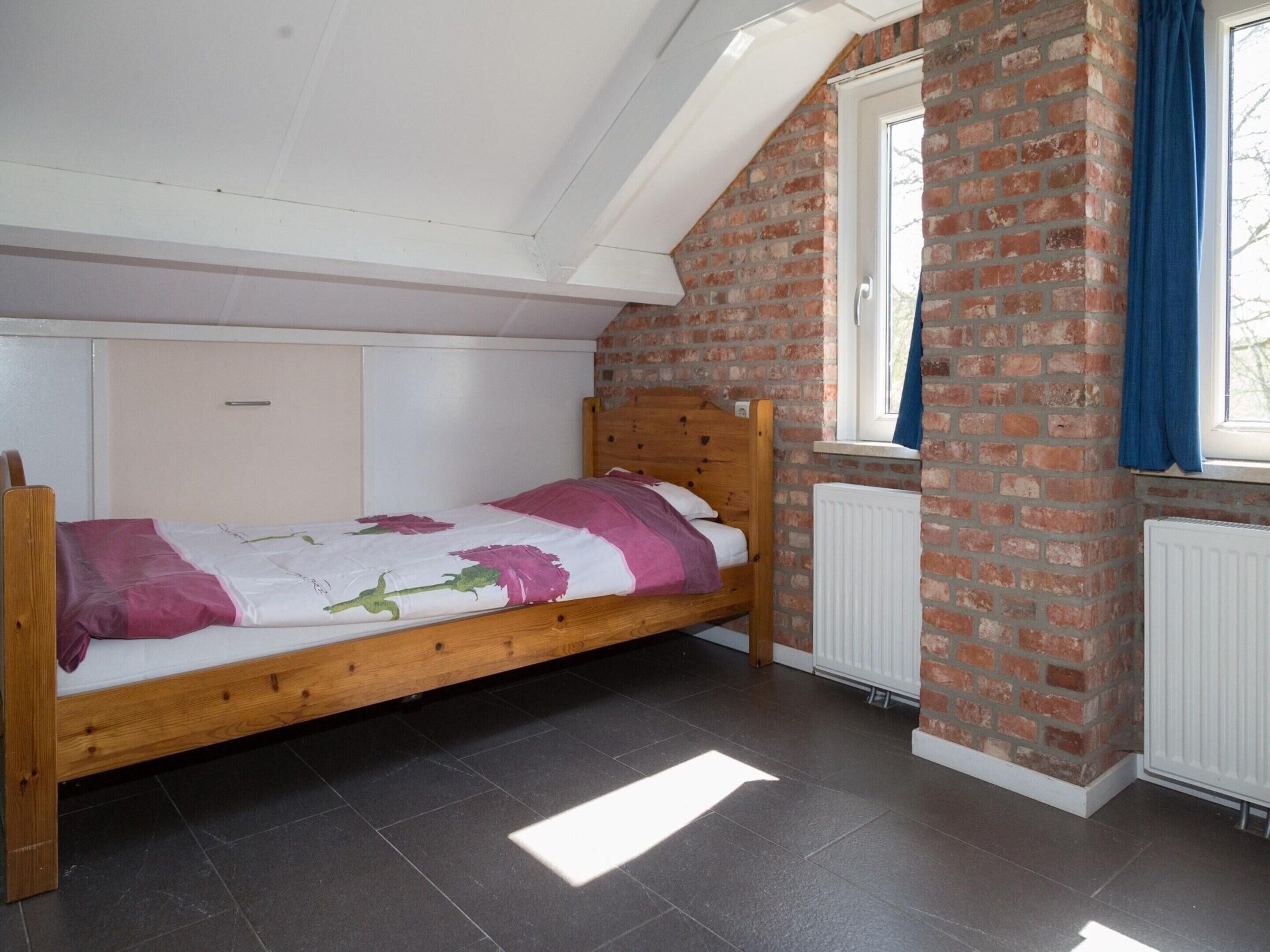 A single bed in a room with a sloped ceiling, brick walls, and a window with blue curtains.