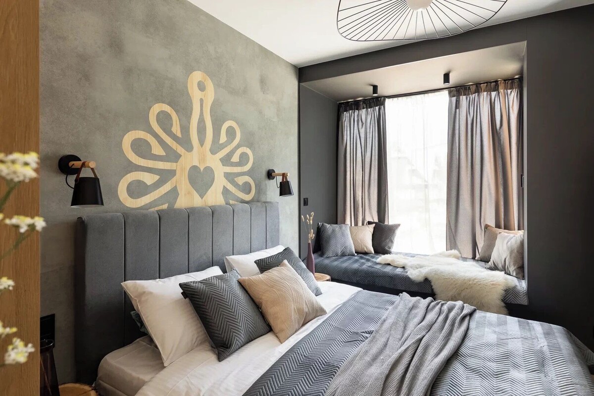 A modern bedroom with a large bed, a smaller sofa, a decorative wall piece, and a window with curtains.