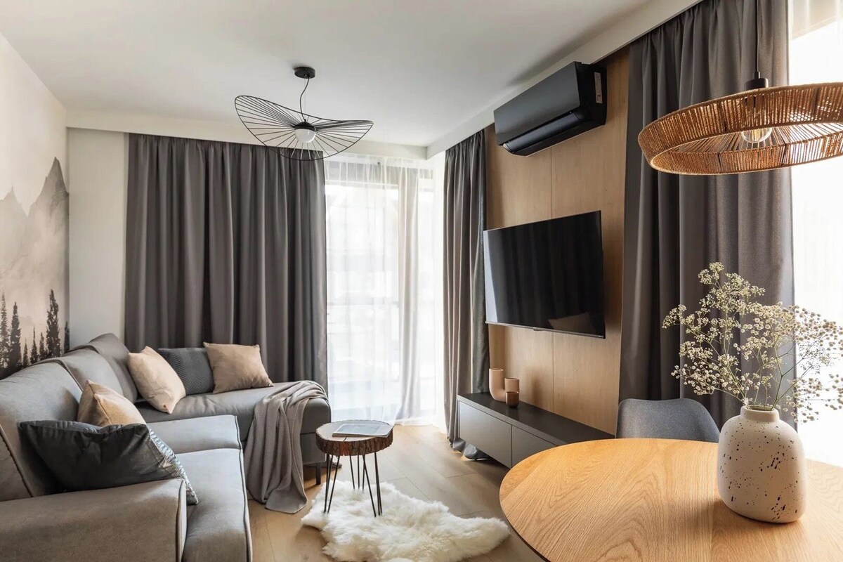 A modern living room with a sofa, a round wooden table, a white furry rug, a wall-mounted TV, and a ceiling fan.