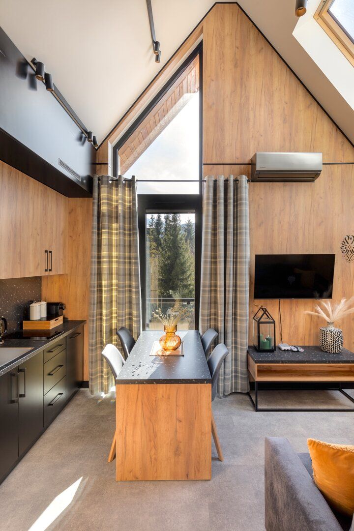 A modern kitchen with a dining area, a flat-screen TV mounted on the wall, and a large window providing a view of the outdoors.