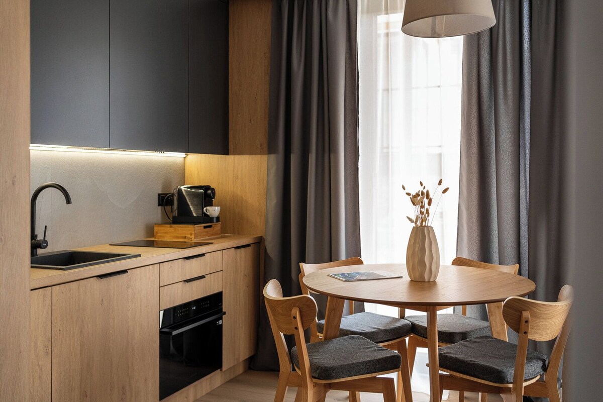 A modern kitchen with a built-in oven, a coffee machine, and a sink. There is a dining table with chairs and a vase with dried plants.