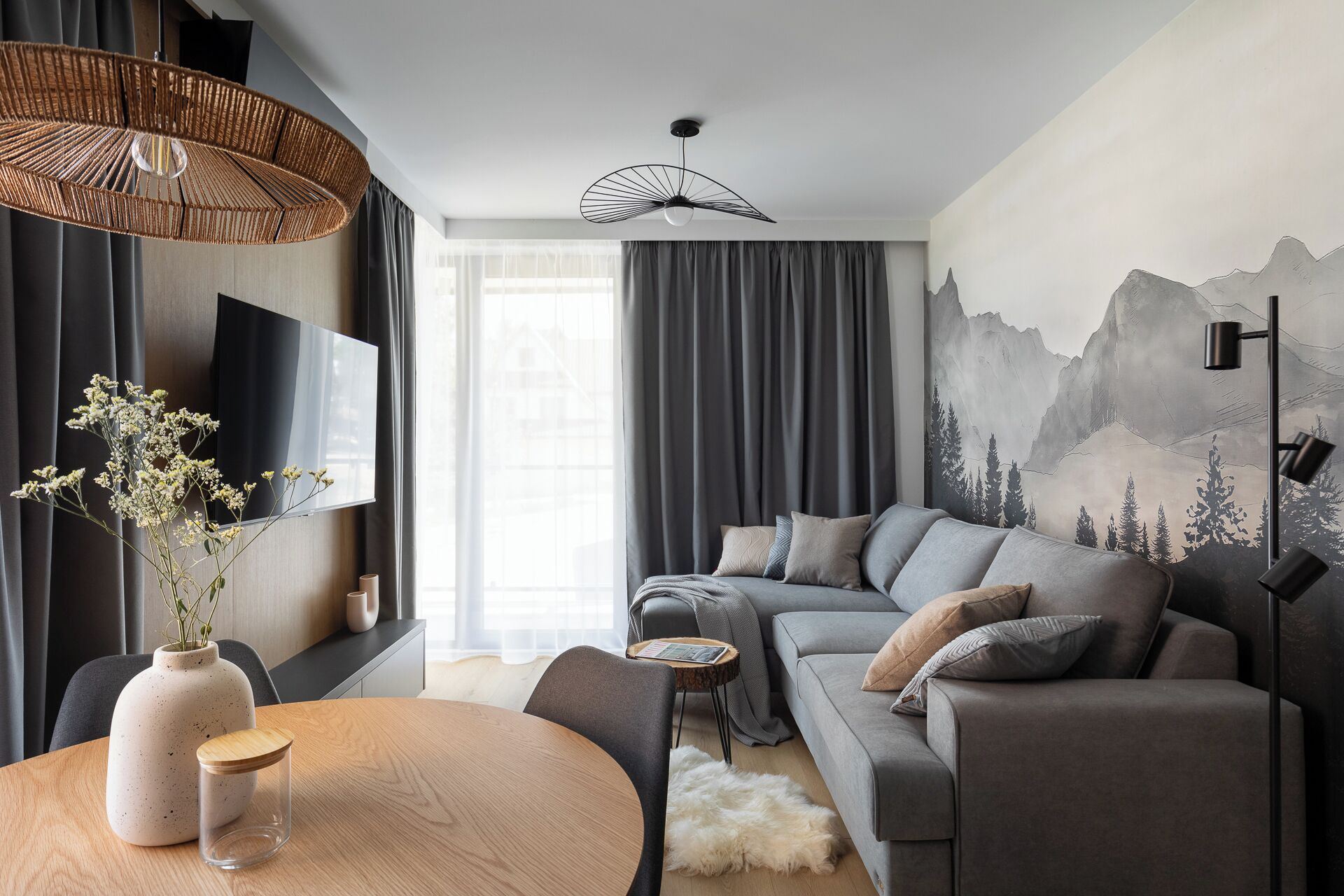 Una sala de estar moderna con un sofá gris, una mesa redonda de madera, un jarrón con flores y un televisor montado en la pared.