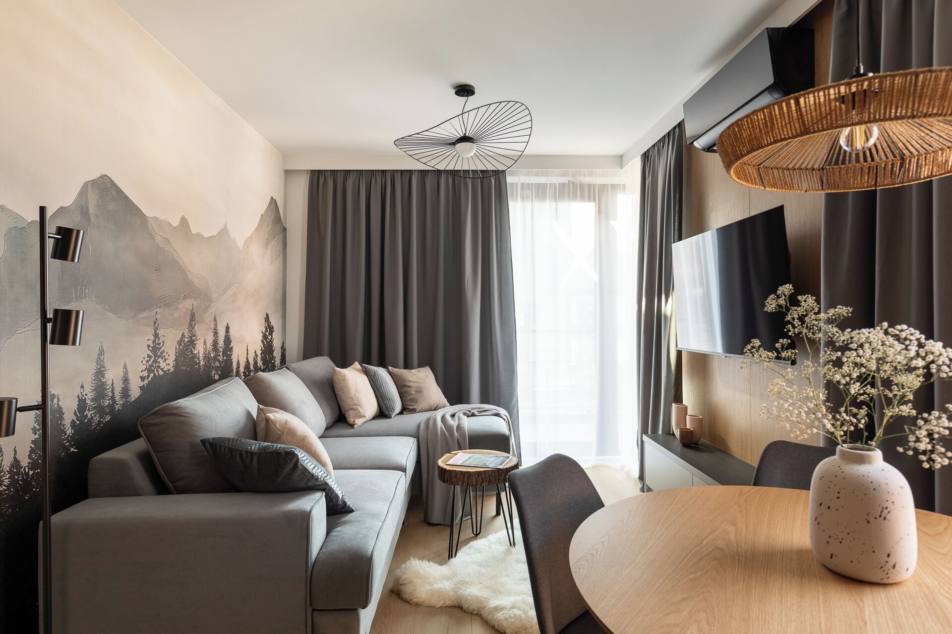 A modern living room with a grey sofa, a round wooden table, and a wall-mounted TV.