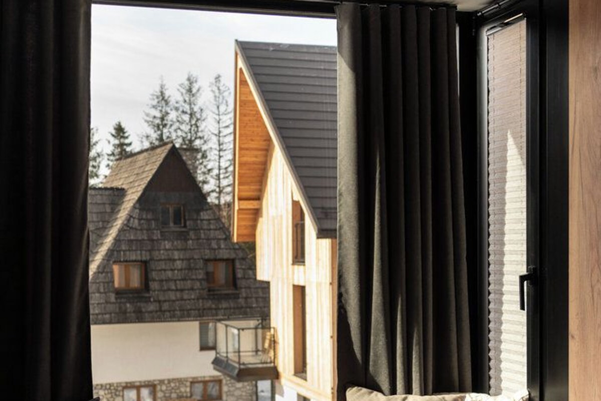 A bedroom with a large window, a bed with patterned bedding, and a view of a wooden house outside.
