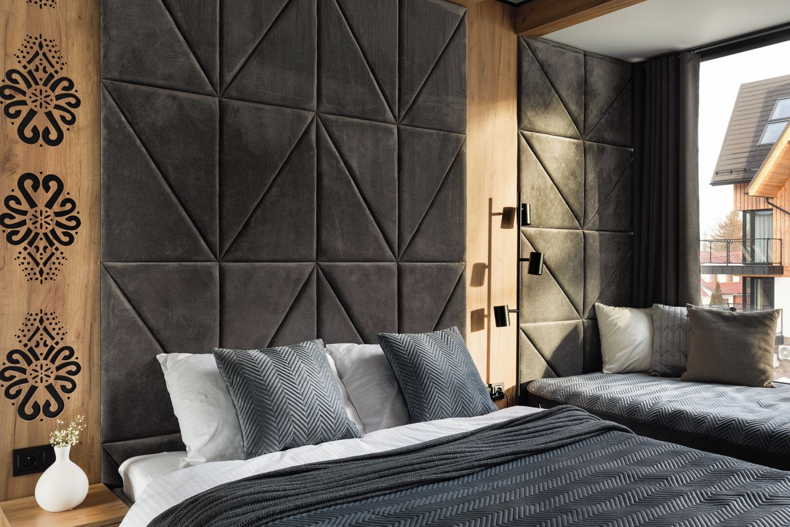 Une chambre moderne avec une tête de lit à motifs géométriques, deux lits avec des oreillers assortis, une table de chevet avec un vase, et une vue sur un immeuble par la fenêtre.