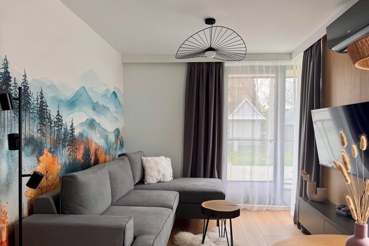 A modern living room with a gray sectional sofa, a round wooden side table, a white furry rug, and a wall-mounted fan.